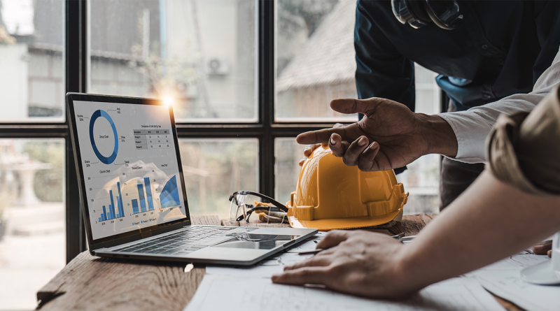 Image of two men stood at a desk with a laptop on displaying different graphs on the screen to support construction project management tools article